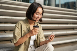 Mulher asiática sentada em uma escada ao ar livre, segurando um café em uma mão e um celular na outra, enquanto olha para a tela.